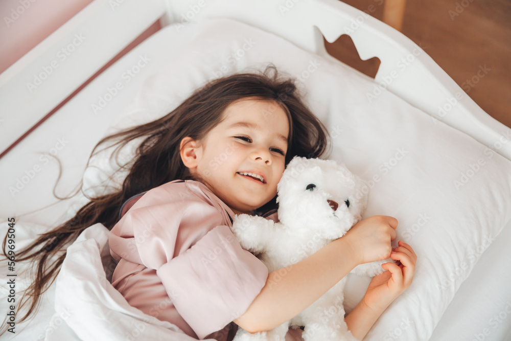 a little dark-haired girl is sleeping in bed, hugging a teddy bear under a white blanket.the child is in bed in the morning.