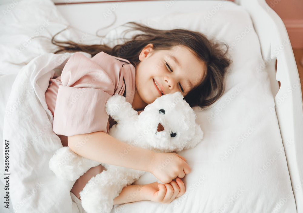 a little dark-haired girl is sleeping in bed, hugging a teddy bear under a white blanket.the child is in bed in the morning. a little dark-haired girl is sleeping in bed, hugging a teddy bear under a white blanket.the child is in bed in the morning.