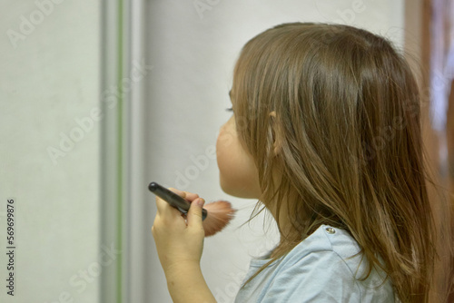 A little girl holds a blush brush to apply powder on her cheeks and face, happily looking in the mirror. Cute baby is playing makeup makeup game..