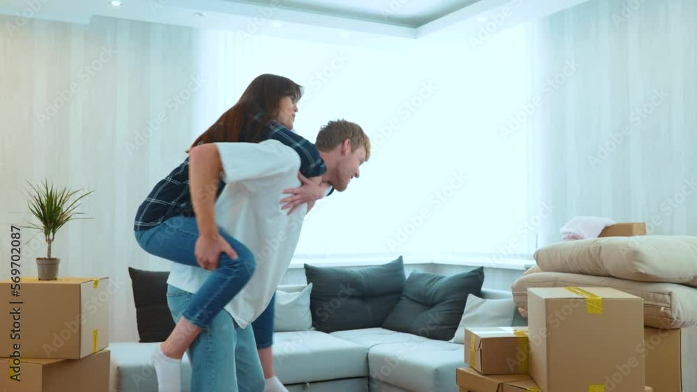 Close up of cheerful Caucasian man and woman having fun playing in new house on moving day. Piggyback ride. Happy young couple in room with many boxes moving in together. Home concept. Real time Close up of cheerful Caucasian man and woman having fun playing in new house on moving day. Piggyback ride. Happy young couple in room with many boxes moving in together. Home concept. Real time