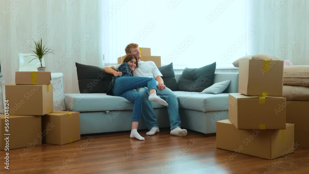 Caucasian joyful boyfriend and girlfriend moving in together in new house. Tired happy young married couple sitting down on sofa in room with many boxes on moving day. Home concept. Real time Caucasian joyful boyfriend and girlfriend moving in together in new house. Tired happy young married couple sitting down on sofa in room with many boxes on moving day. Home concept. Real time