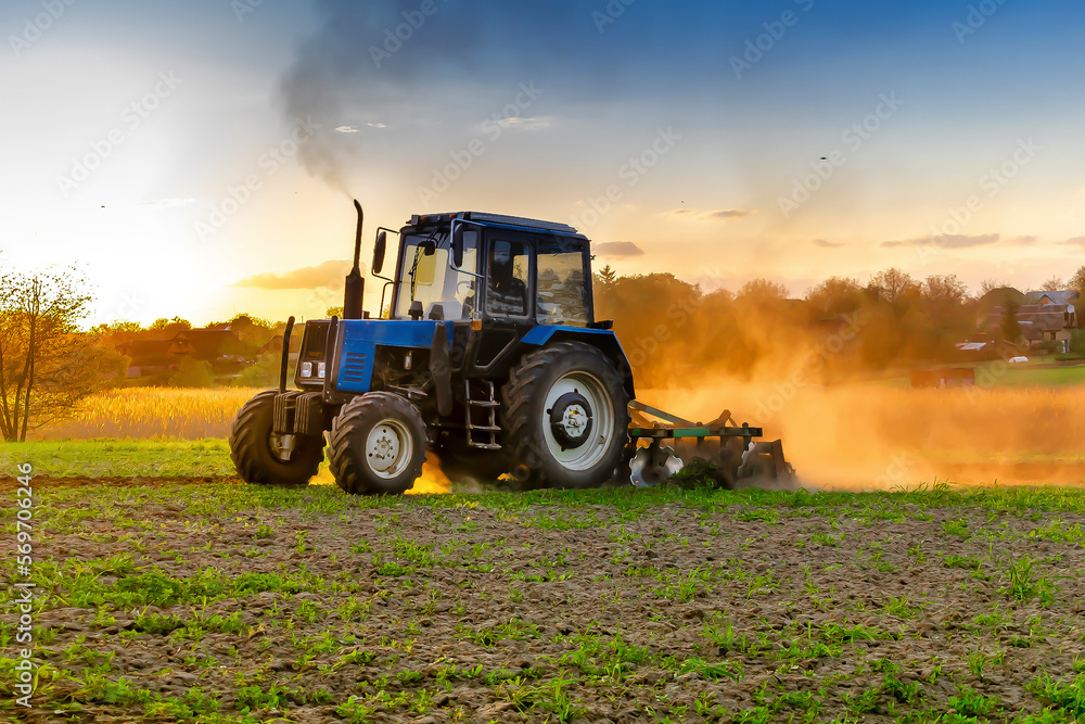 Modern blue tractor machinery plowing agricultural field meadow at farm at spring autumn during sunset.Farmer cultivating,make soil tillage before seeding plants,crops,nature countryside rural scene