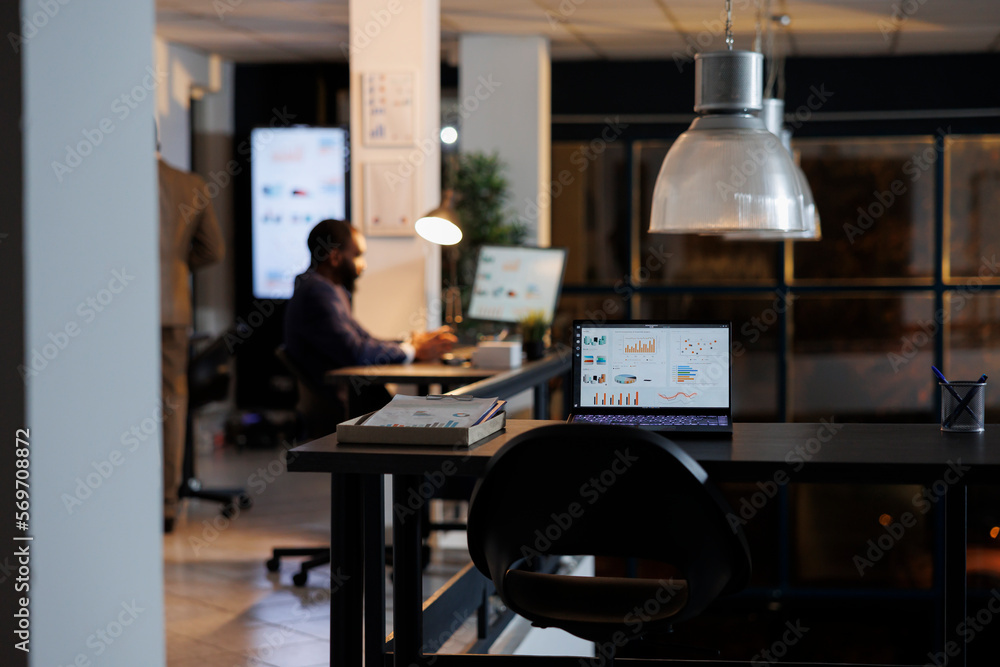 Laptop computer with company investment graph on screen, standing on desk in startup office. In background businessman analyzing marketing statistics, typing strategy. Business concept Laptop computer with company investment graph on screen, standing on desk in startup office. In background businessman analyzing marketing statistics, typing strategy. Business concept