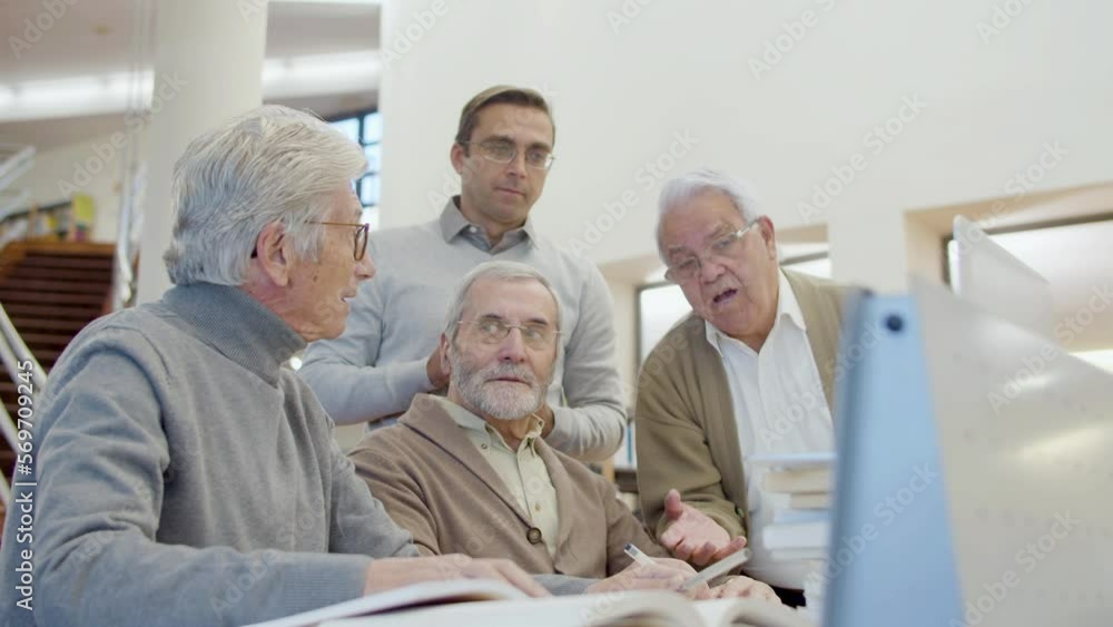 Senior men and teacher looking laptop screen in library. Elderly friends sitting at table, having class with teacher, working together, analyzing information. Education, retirement concept Senior men and teacher looking laptop screen in library. Elderly friends sitting at table, having class with teacher, working together, analyzing information. Education, retirement concept