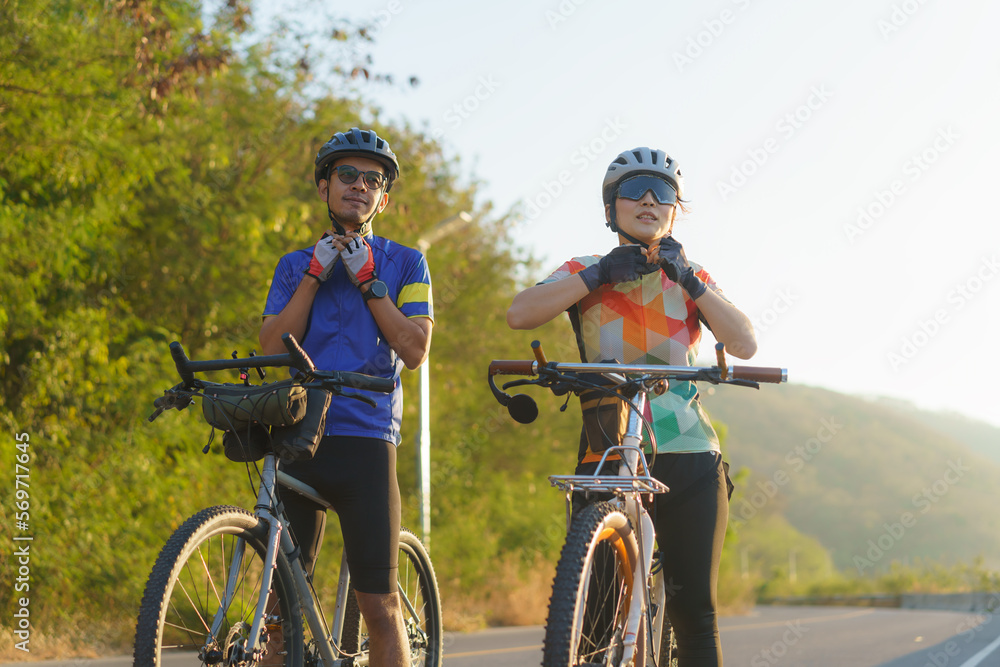 Asian cyclist couple dons helmets and chin straps as they prepare for their bike ride around a beautiful morning lake.