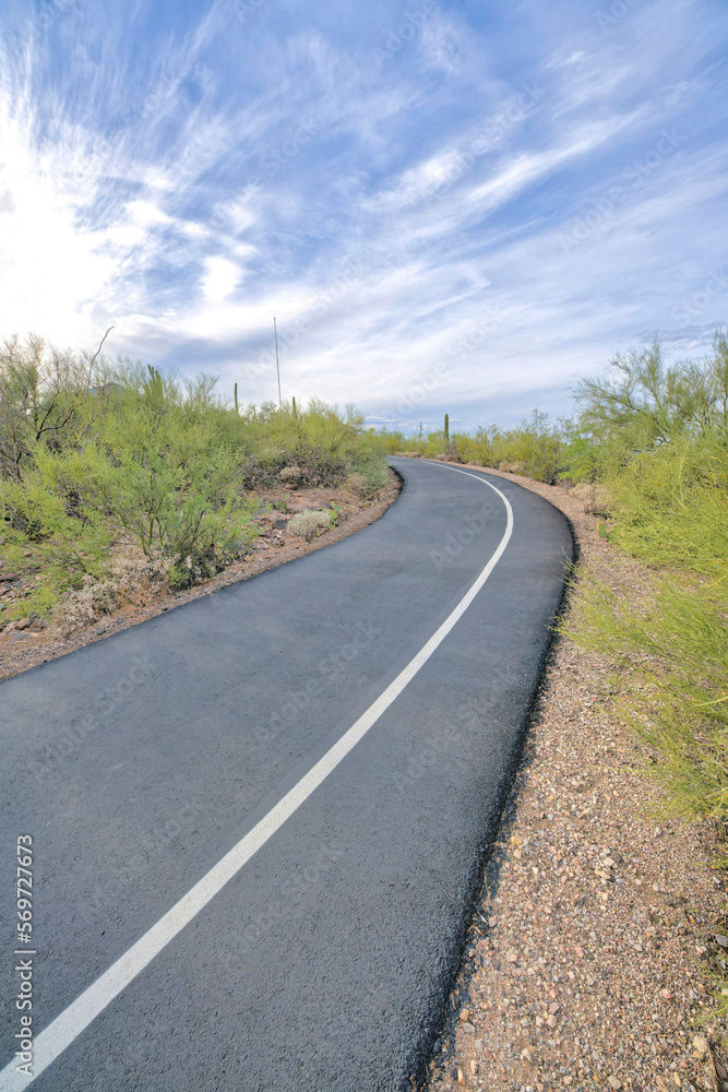Uphill asphalt bike path and walking pathway at Tucson, Arizona. Pathway with white line on the right in the middle of desert land and slope against the sky background.