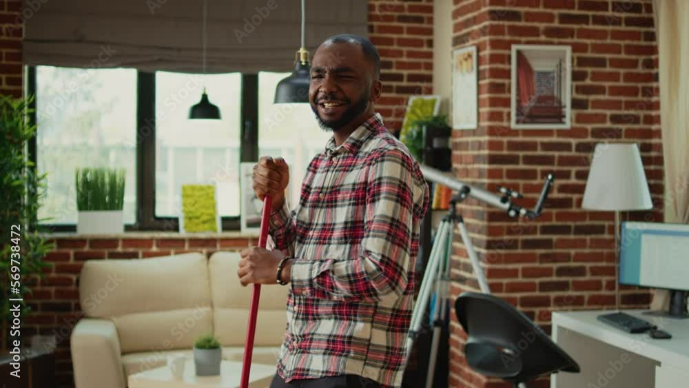Happy smiling adult mopping living room floor to clean dirt, using mop and all purpose cleaner at home. Young person dancing and listening to music while sweeping dust or mess. Happy smiling adult mopping living room floor to clean dirt, using mop and all purpose cleaner at home. Young person dancing and listening to music while sweeping dust or mess.