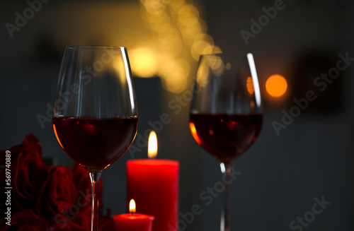 Glasses of red wine, burning candles and rose flowers against blurred lights. Romantic atmosphere