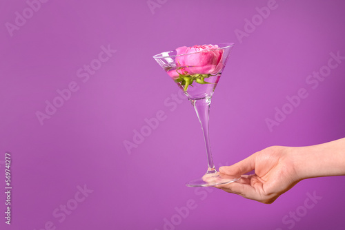 Woman holding martini glass with water and pink flowers on violet background, closeup. Space for text