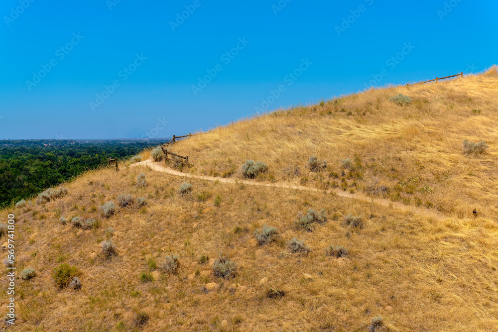 Hiking trail in the middle of a mountain slope in Boise, Idaho. Mountain trail on a slope with views of trees on the left against the clear blue skies.