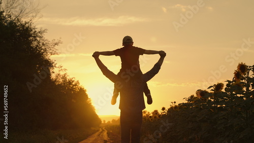 Happy child, father play in field of ripening sunflowers. Little son on shoulders of his father. Boy and dad are traveling across field. Child, parents play in nature. Happy family, childhood concept
