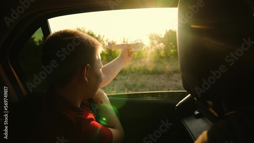 Boy dreams of flying, adventures, looks out open window of car. Happy family enjoy car travel outdoors together. Childrens journey, Little boy is sitting in car playing with wooden toy airplane