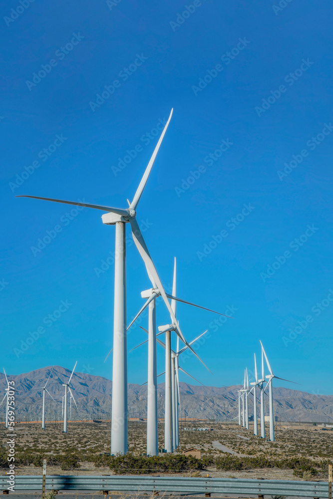 Windmills on a wind farm for generation of green energy on a sunny day. High wind turbines or windpumps on a wind field with mountains and clear blue sky views. Windmills on a wind farm for generation of green energy on a sunny day. High wind turbines or windpumps on a wind field with mountains and clear blue sky views.