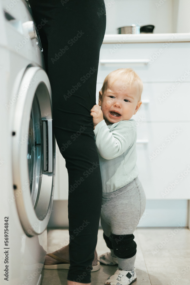 Little infant baby boy child hiding between mothers legs demanding her attention while she is multitasking, trying to do some household chores in kitchen at home. Mother on maternity leave Little infant baby boy child hiding between mothers legs demanding her attention while she is multitasking, trying to do some household chores in kitchen at home. Mother on maternity leave