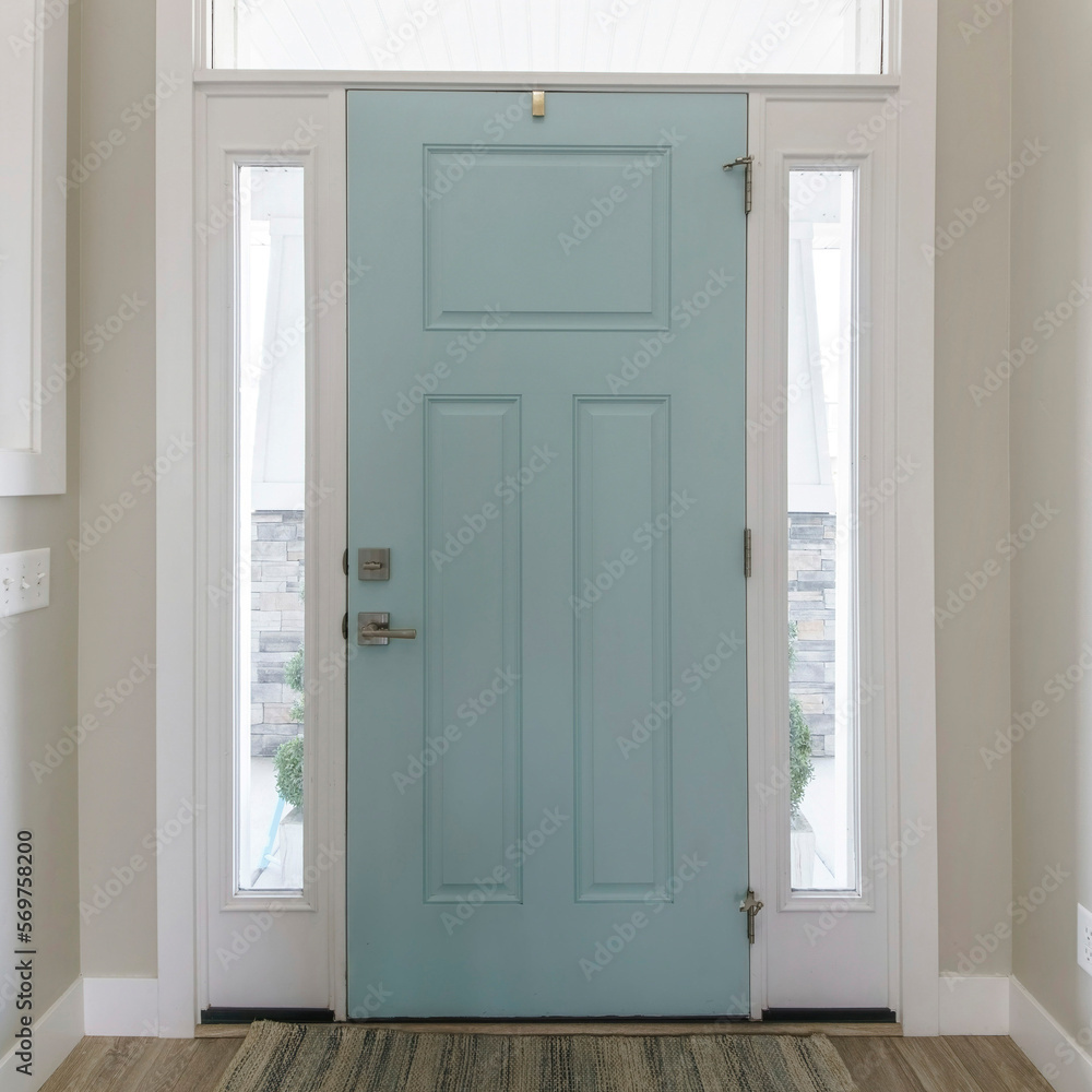 Square Mint front door interior with transom window and sidelights. Interior of a house with light gray walls, wooden flooring and a view of two rooms on both sides with windows.