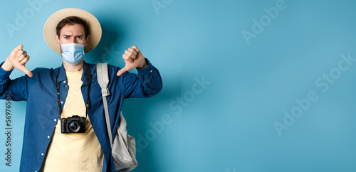 Covid-19 and summer holidays concept. Disappointed tourist in medicla mask frowning, showing thumbs down, negative feedback about travel, standing on blue background photo