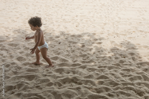 Cute boy toddler on summer vacation at the beach. He has brown curly hair and is wearing a diaper. He is exploring and walking on the sand for the first time.