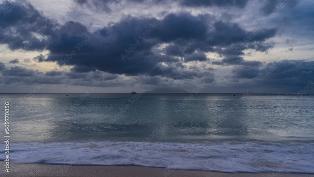 Obraz premium Evening seascape in shades of blue. Picturesque clouds in the sky. Yachts and the silhouette of the island on the horizon. Glare on the ocean surface. Foam of waves on a sandy beach. Long exposure