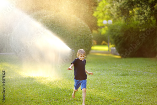 Funny little boy playing with garden sprinkler in sunny city park. Elementary school child laughing  jumping and having fun with spray of water. Summer outdoors activity for kids.