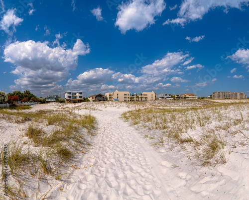 White sand path in between dunes with grasses on a beach near Noriego Point in Destin, Florida. Path with footprints near dunes heading to the residential buildings under the sky background.