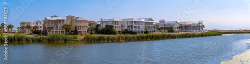 Lakefront homes on the beach at Destin Point, Destin, Florida panorama. There is a lake at the front with tall grasses on the shore near the three-storey houses against the bright sky background.