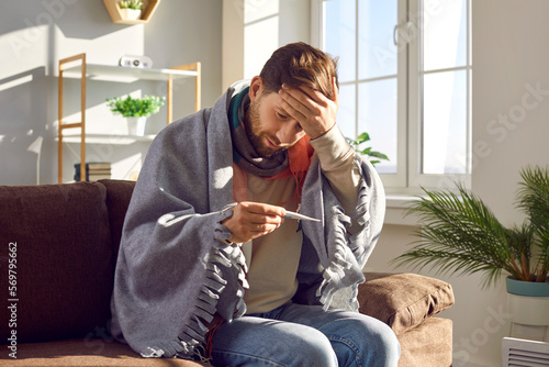 Fotografia Sick young man sitting on sofa and checking his temperature wrapped in blanket and touching his forehead with hand