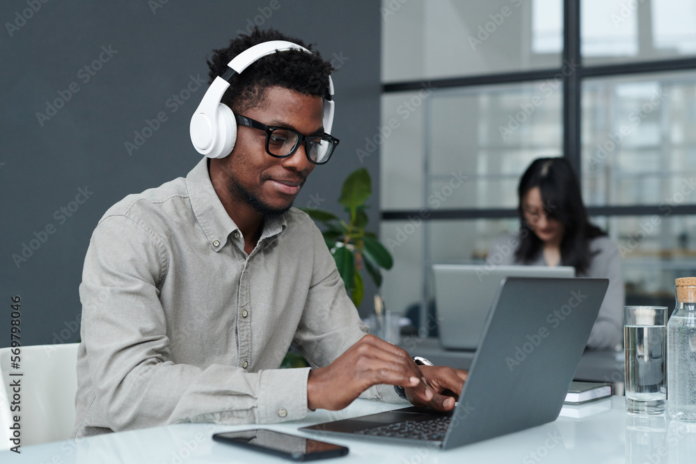 Young African American businessman in wireless headphones typing on laptop during his work at table in office - obrazy, fototapety, plakaty 