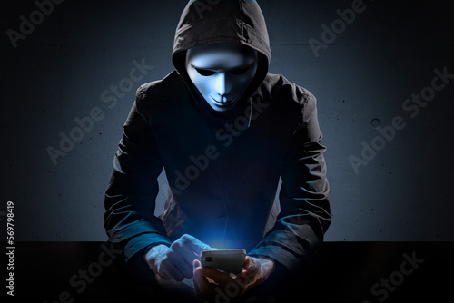 Foto 仮面を被り、スマートフォンを操作している犯罪者。
ハッカー、ネット中傷などのイメージ