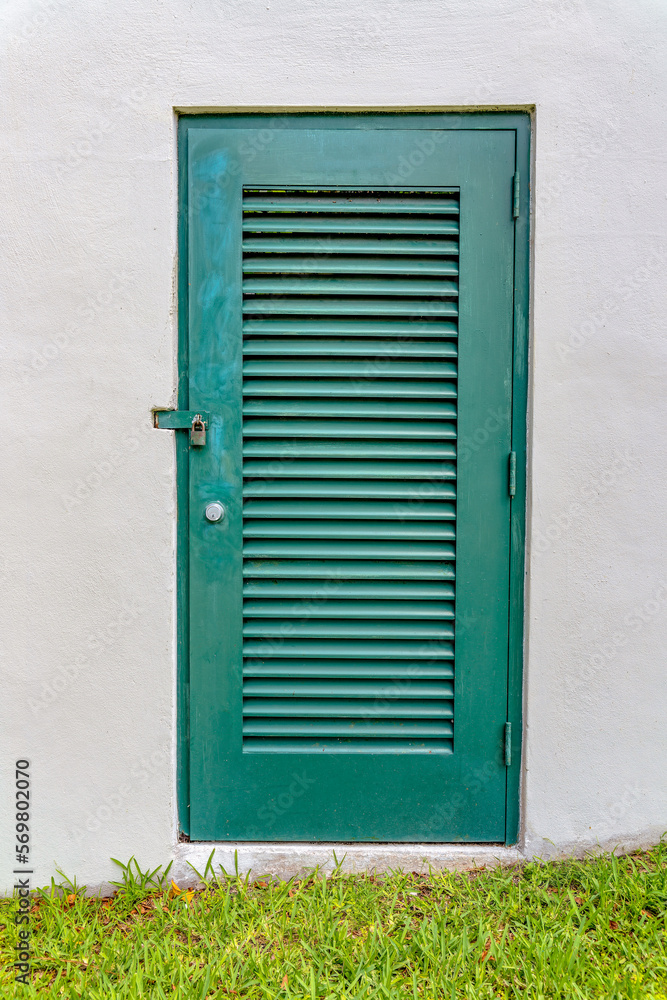Green louvered door of a hidden electrical utility room of an irrigation at Miami, Florida. Door with padlock on a building with white wall and lawn outdoors.
