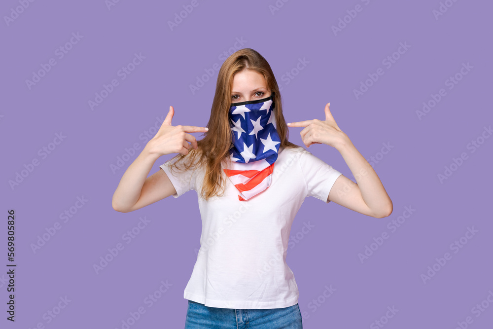 Young woman with mask in form flag america on her face in white t-shirt shows a gesture with her fingers at herself while standing on a purple background