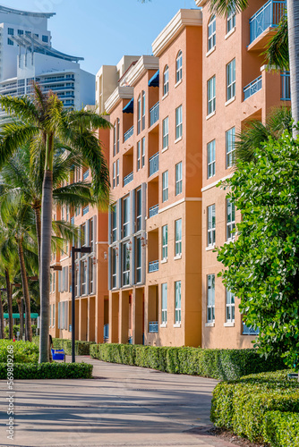 Concrete path with bushes and palm trees at front of an orange apartment building- Miami, Florida. Pathway heading to the front of a multi-storey building with balconies.