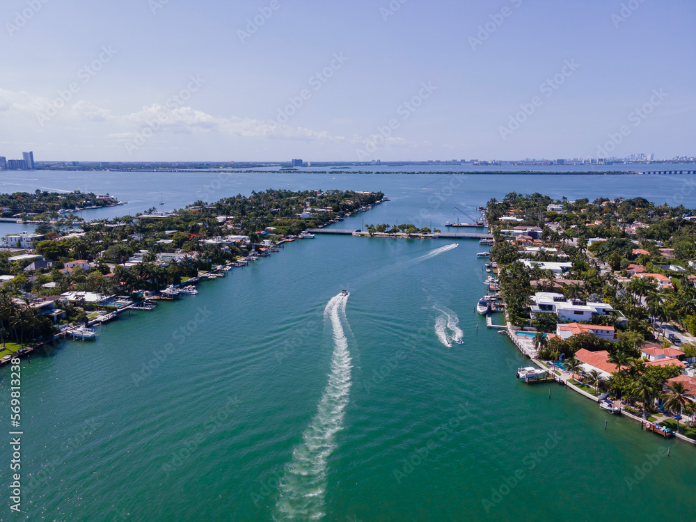Waterfront homes in the middle of the Intracoastal Waterway in Miami Florida. Aerial view of a man-made inland water channel with houses, boats, and trees against blue sky.