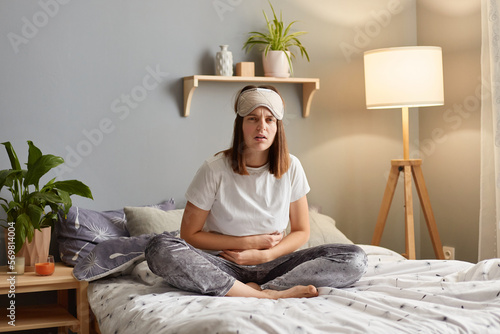 Portrait of upset Caucasian woman suffering from strong abdominal pain, touching her tummy, sitting on bed, feeling acute stomachache, has chronic disease, spasm, or hernia. photo