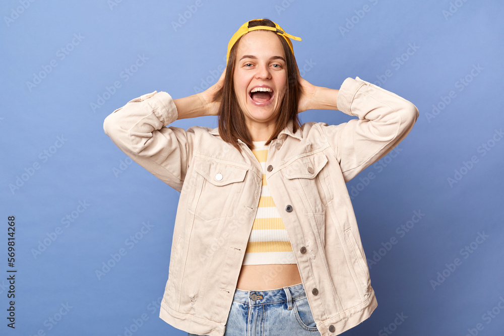 Photo of extremely happy cheerful satisfied brown haired teenager girl in baseball cap and jacket, keeps hands behind her head, looking at camera and laughing, posing isolated over blue background. Photo of extremely happy cheerful satisfied brown haired teenager girl in baseball cap and jacket, keeps hands behind her head, looking at camera and laughing, posing isolated over blue background.