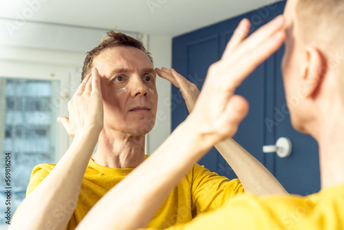 Middle aged man apply cream on clear face, then massage with hands. Male portrait in room mirror. Skincare morning routine, careful beauty treatment, home lifestyle. Facial care concept.