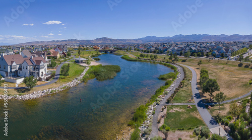 Panorama aerial view of Daybreak community in Utah with lake in the middle with pathways on the side. There are houses surrounding the lake with grassy shore and pathways on the side.
