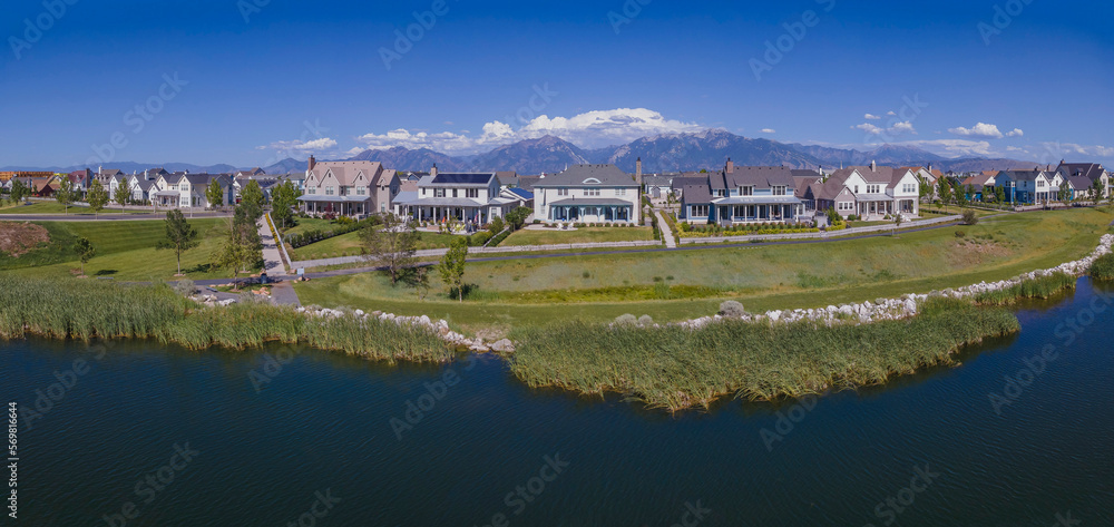 Panorama of residential neighborhood in Daybreak Utah overlooking a river. Facade of houses with towering mountains and blue sky background on a sunny day.