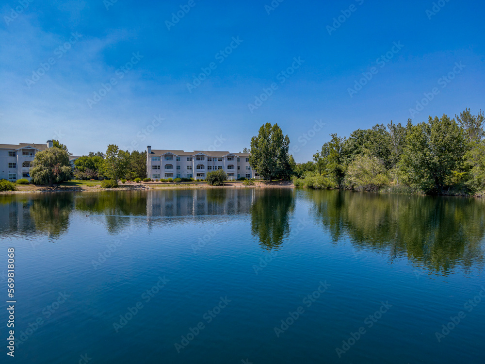 Apartments with beautiful view of a reflective small lake in Boise Idaho. The lush green trees are reflected on the calm water surface with bright blue sky overhead on this sunny day. Apartments with beautiful view of a reflective small lake in Boise Idaho. The lush green trees are reflected on the calm water surface with bright blue sky overhead on this sunny day.