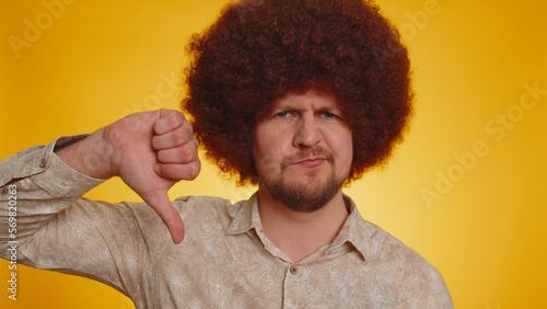 Dislike. Upset unhappy man with lush Afro hairstyle coiffure showing thumbs down sign gesture, expressing discontent, disapproval, dissatisfied, dislike. Young guy. Indoor studio on yellow background © Andrii Iemelianenko