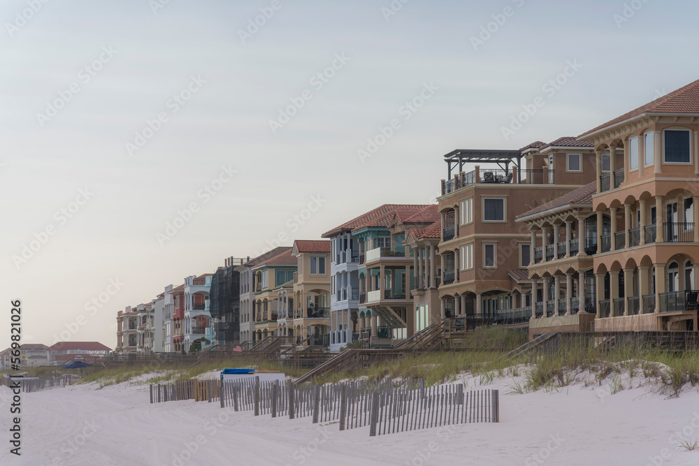 Row of villas and beach houses with wooden bridges over sand dunes in Destin, Florida. There are lined wood fences at the front near the sand dunes with grasses.