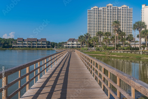 Boardwalk over the water with views of buildings at Destin, Florida. Wooden boardwalk with railings against the view of three-storey and multi-storey buildings with blue sky background. © Jason