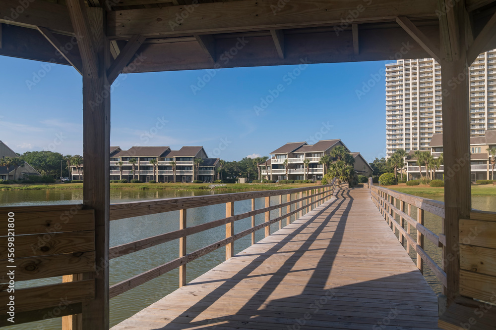 Views of a wooden boardwalk heading to residences under a gazebo in Destin, Florida. Views under the roof of a gazebo connected to a boardwalk over the lake heading to the apartments at the back.