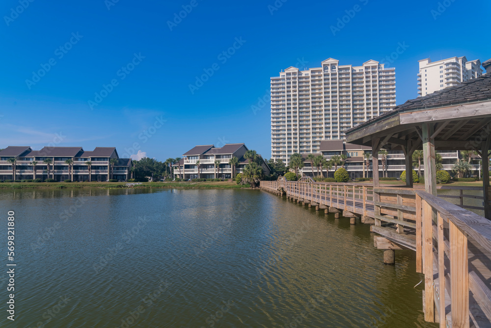 Residential area with waterfront views and boardwalk in Destin, Florida. There is a boardwalk with gazebo on the right over the water against the modern apartments and blue sky at the background.