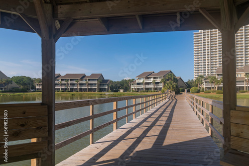 Views of a wooden boardwalk heading to residences under a gazebo in Destin, Florida. Views under the roof of a gazebo connected to a boardwalk over the lake heading to the apartments at the back. © Jason