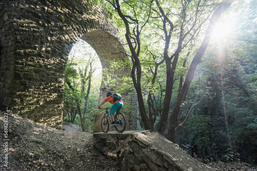 Man riding bike over footpath by arched stone wall in forest