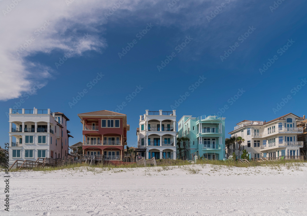 Beach houses facade with balconies and roof decks facing the white sand shore in Destin, Florida. There are footbridges as beach access over the sand dunes at the front of the colorful beach houses.