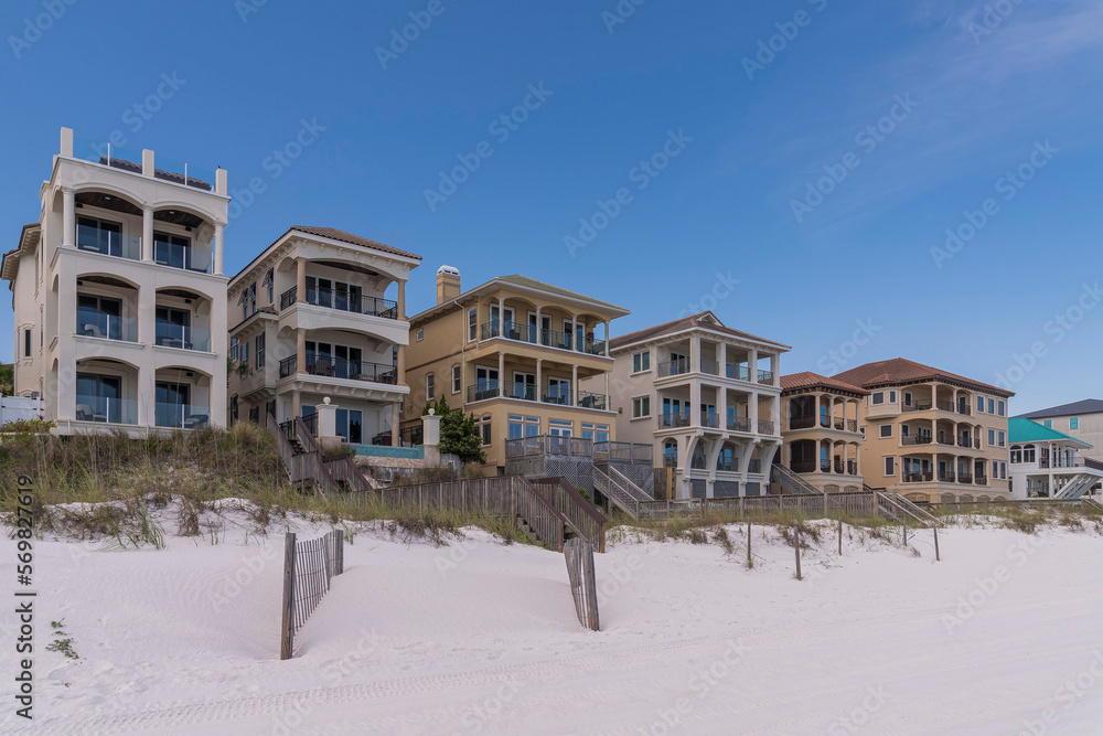 Three-storey beach houses with fenced grassy sand dunes at Destin, Florida. Facade of buildings with balconies and footbridges over the grassy dunes against the blue sky.