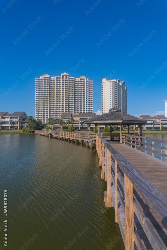 Vertical shot of wooden footbridge over Stewart Lake in Destin, Florida. Walkway with railings heading to the apartments and hotels with low-rise and high-rise structures against blue sky.