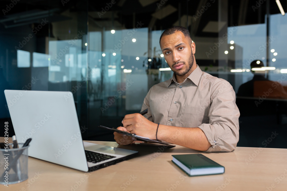 Portrait of serious and focused businessman, man looking thinking at camera writing and signing document inside office, african american financier at work on laptop.