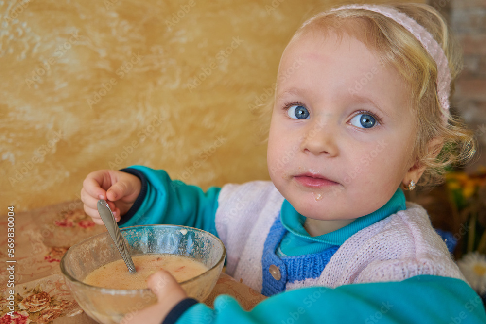 Naklejka premium little girl eats porridge,a beautiful child at the table with a bowl of porridge, beautiful blue eyes
